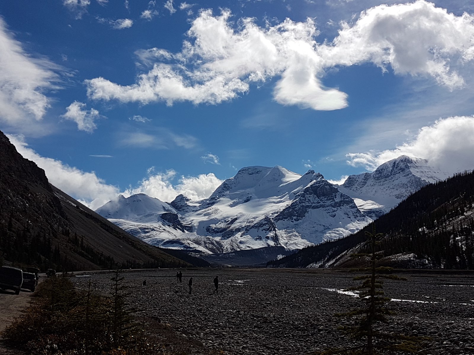 Snow-covered mountains beneath a blue sky and white clouds, framed by forested slopes.